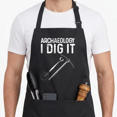 Archaeology artifact apron - White distressed text ARCHAEOLOGY I DIG IT above crossed shovel and pickaxe tools — shown on a black cotton canvas apron against a white background