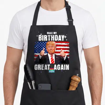 Trump tea apron - Make My Birthday Great Again text around Trump thumbs up and small cake on flag background — shown on a black cotton canvas apron against a white background