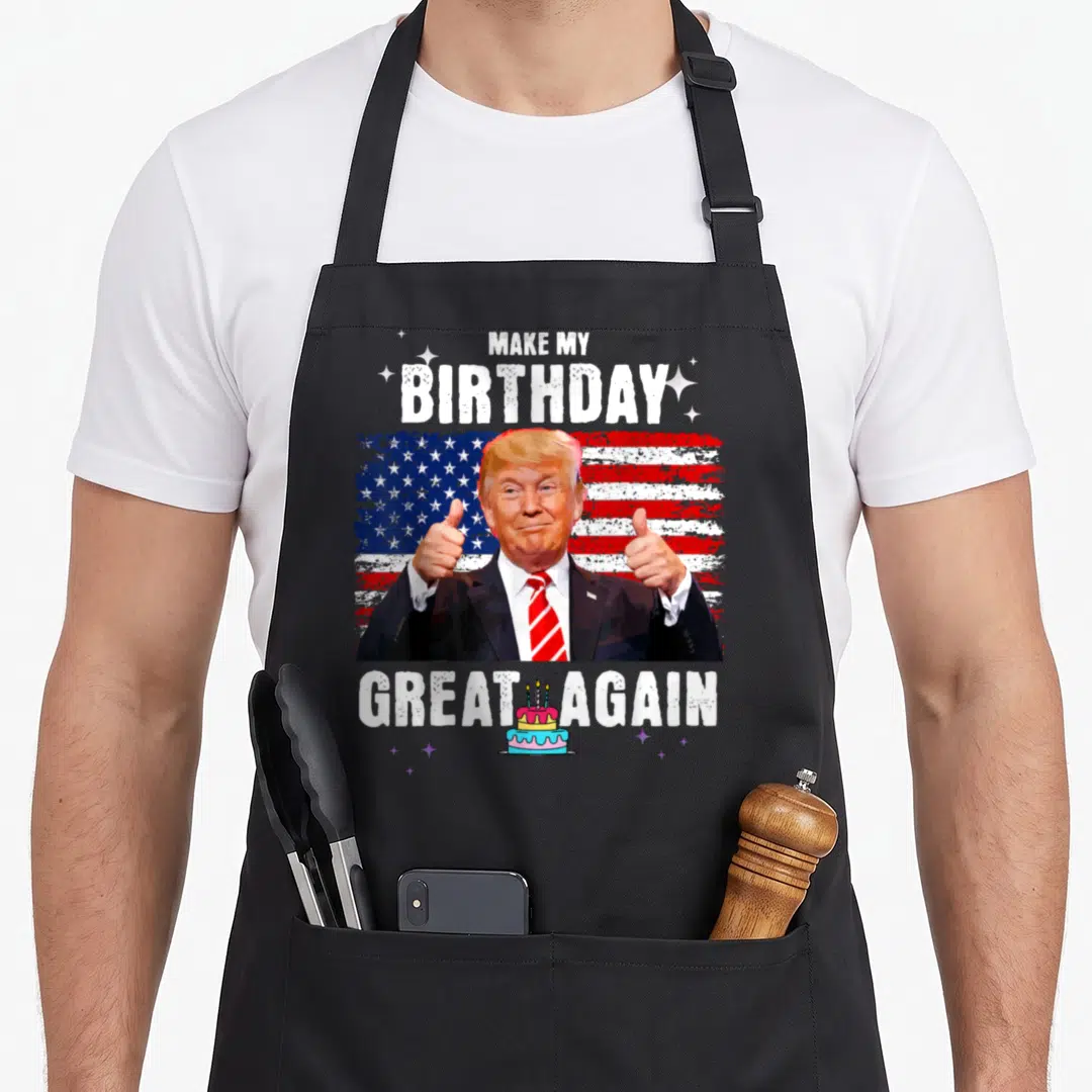 Trump tea apron - Make My Birthday Great Again text around Trump thumbs up and small cake on flag background — shown on a black cotton canvas apron against a white background