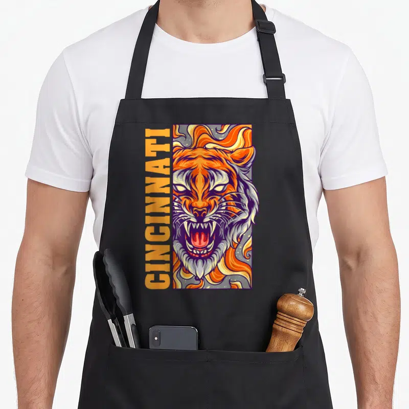 Growling bengal cat apron - Vertical Cincinnati text beside orange tiger head with purple outlines and yellow flame — shown on a black cotton canvas apron against a white background