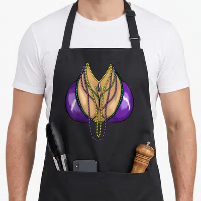 Gras fat chef apron - Cartoon cleavage framed by purple shapes draped with three colorful bead strings — shown on a black cotton canvas apron against a white background