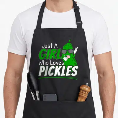 Meme pickle apron - Cartoon pickle wearing sunglasses gesturing next to stacked text Just A GIRL Who Loves PICKLES — shown on a black cotton canvas apron against a white background