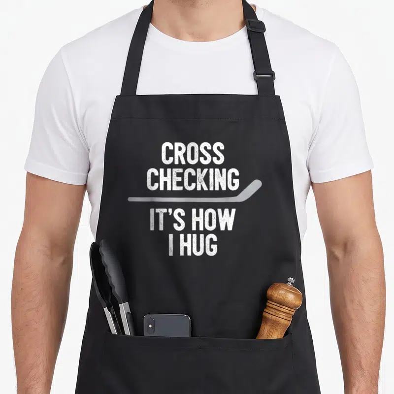 Hockey cross apron - CROSS CHECKING IT'S HOW I HUG text with horizontal hockey stick in distressed white — shown on a black cotton canvas apron against a white background