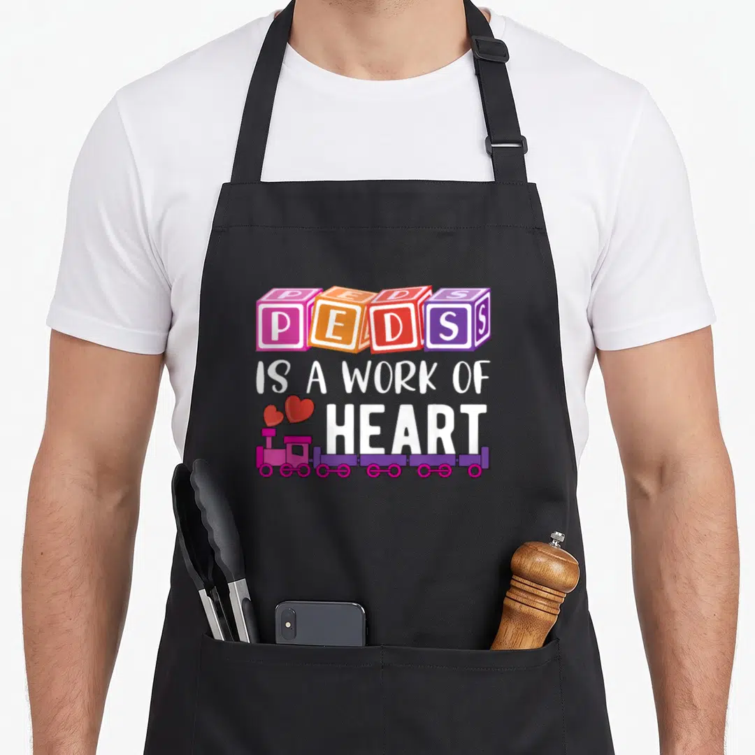 Pediatrician nurse apron - Four colorful toy blocks spelling PEDS above white text and a pink train pulling two red — shown on a black cotton canvas apron against a white background