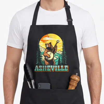 Carolina bear apron - Retro bear playing banjo in front of mountains with ASHEVILLE text below — shown on a black cotton canvas apron against a white background