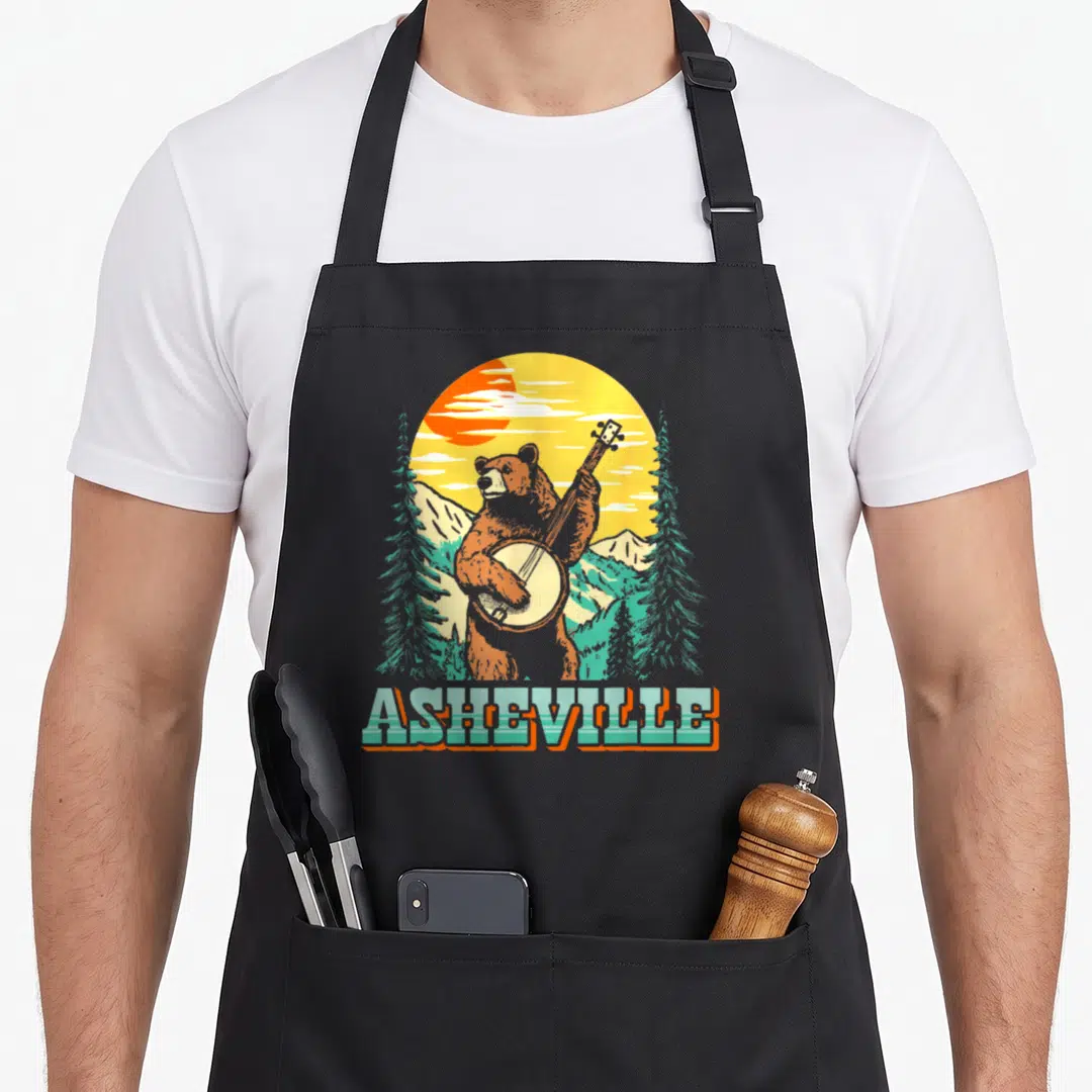 Carolina bear apron - Retro bear playing banjo in front of mountains with ASHEVILLE text below — shown on a black cotton canvas apron against a white background