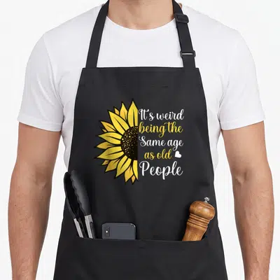 Sunflower weird aprons - Half sunflower with It's weird being same age as old people text — shown on a black cotton canvas apron against a white background