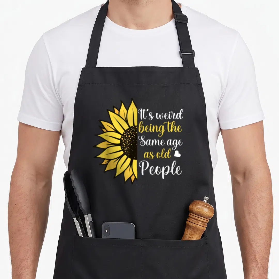 Sunflower weird aprons - Half sunflower with It's weird being same age as old people text — shown on a black cotton canvas apron against a white background