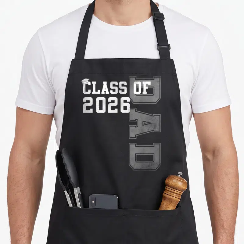 Graduate dad grad apron - White distressed CLASS OF 2026 text with small graduation cap on letter C — shown on a black cotton canvas apron against a white background
