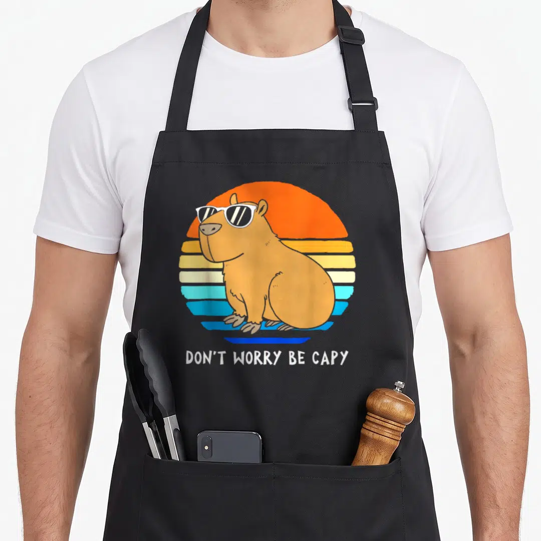 Rodent capybara apron - Brown capybara in white sunglasses before striped orange blue sun with text DON'T WORRY BE CAPY — shown on a black cotton canvas apron against a white background