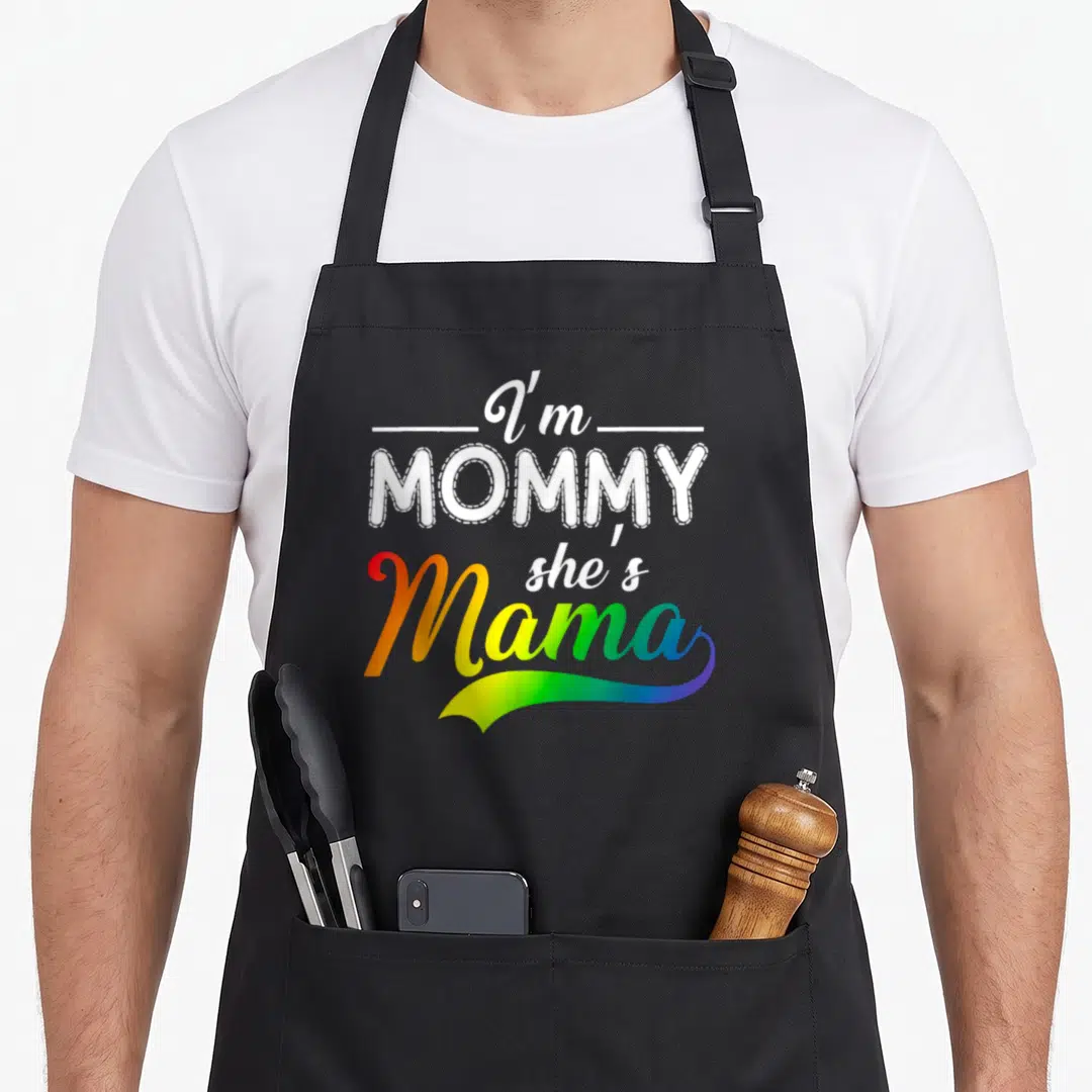 Funny lesbian mama apron - I'm MOMMY she's Mama typography with rainbow gradient and stitched border text — shown on a black cotton canvas apron against a white background