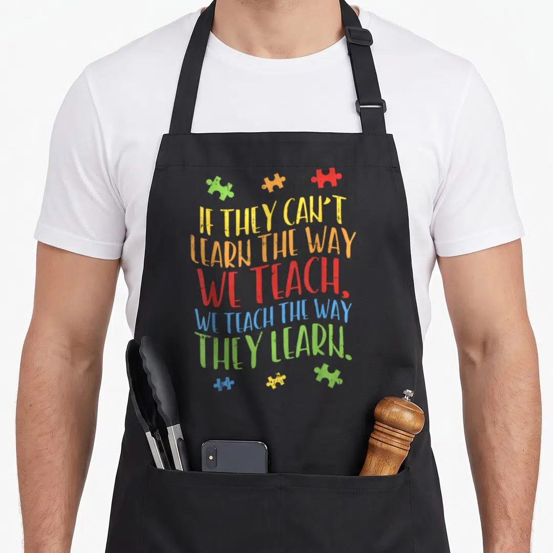 Autistic teacher art apron - Stacked colorful text with six puzzle pieces scattered around the top and bottom — shown on a black cotton canvas apron against a white background