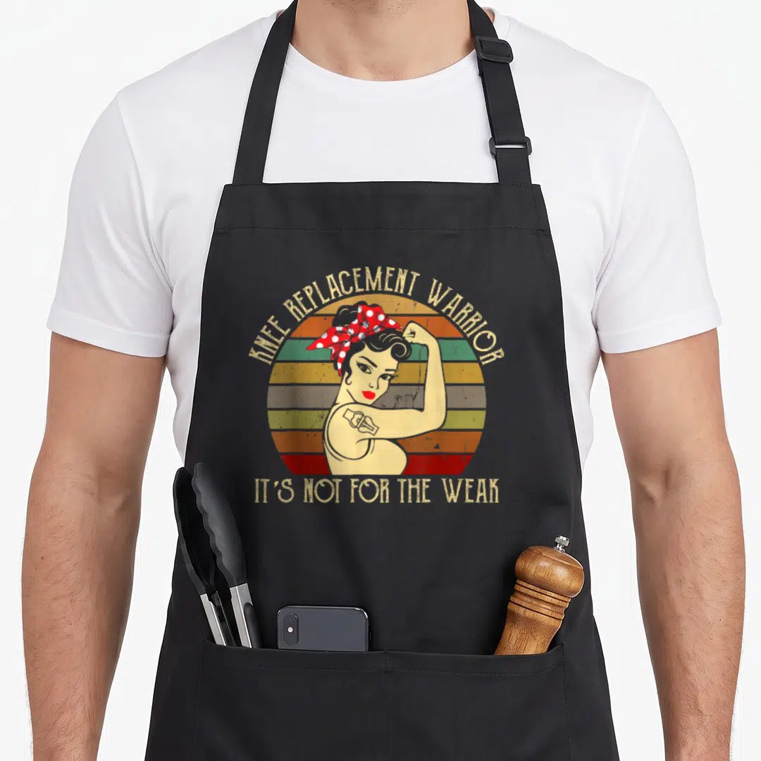 Funny warrior knee replacement apron - Retro woman flexing arm with knee implant on striped sun background — shown on a black cotton canvas apron against a white background