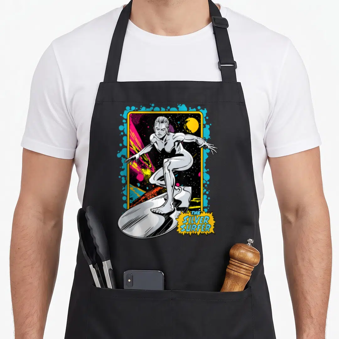 Surfer first steps apron - The Silver Surfer surfing in yellow frame with blue pink black text burst — shown on a black cotton canvas apron against a white background