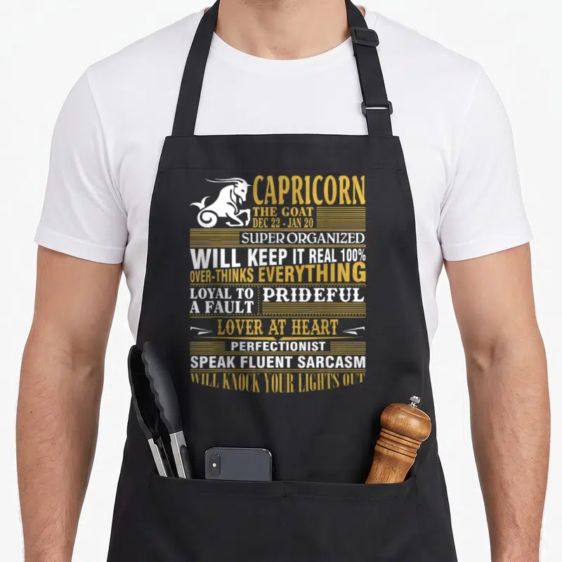 Fact capricorn apron - White goat silhouette next to stacked gold and white text listing zodiac personality traits — shown on a black cotton canvas apron against a white background
