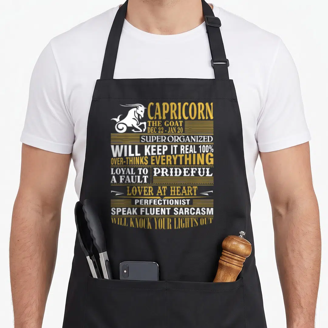 Fact capricorn apron - White goat silhouette next to stacked gold and white text listing zodiac personality traits — shown on a black cotton canvas apron against a white background