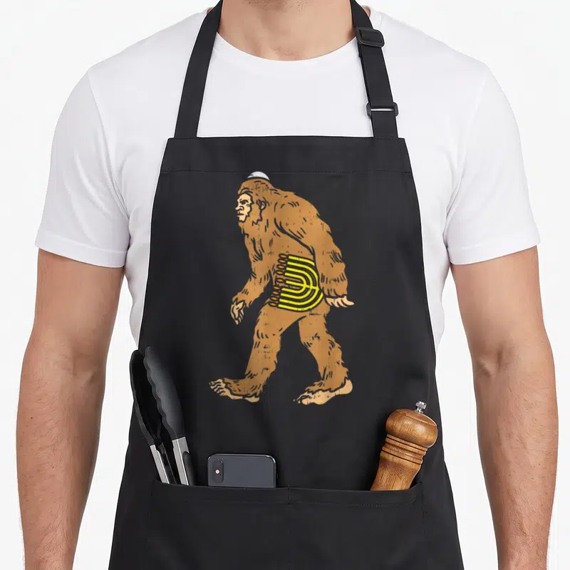 Bigfoot jewish aprons - Cartoon brown bigfoot in profile wearing white kippah and carrying yellow menorah on side — shown on a black cotton canvas apron against a white background