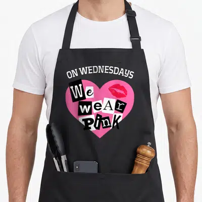 Wednesday valentine apron - ON WEDNESDAYS We wear PINK text in a pink heart with red lips and ransom note letters — shown on a black cotton canvas apron against a white background