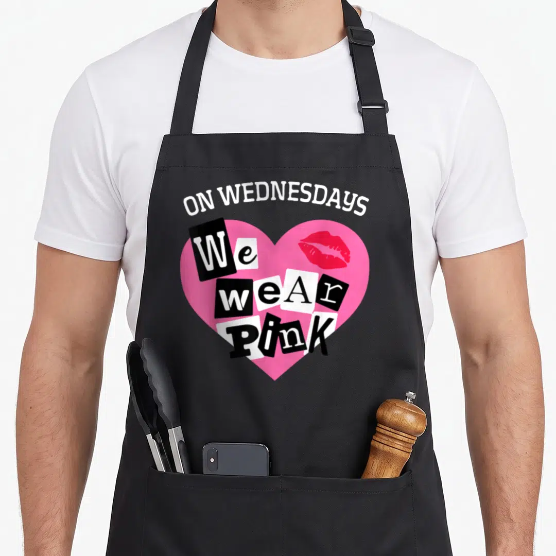 Wednesday valentine apron - ON WEDNESDAYS We wear PINK text in a pink heart with red lips and ransom note letters — shown on a black cotton canvas apron against a white background