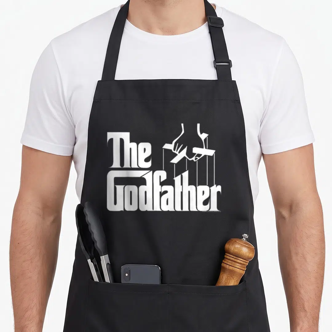 Godfather fathers apron - White puppet hand holding crossbar above The Godfather text in minimalist style — shown on a black cotton canvas apron against a white background