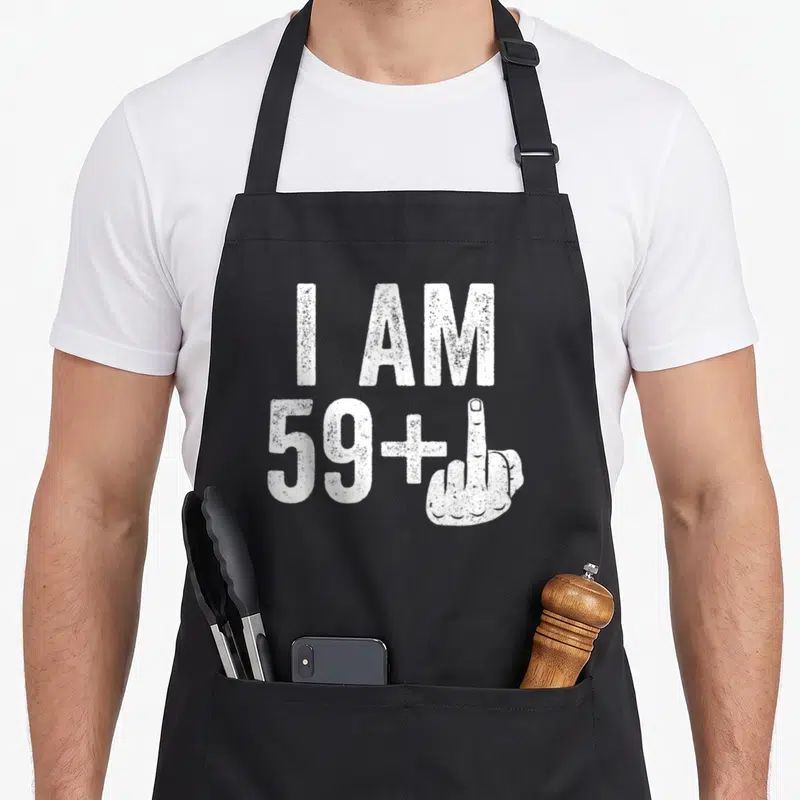 Middle 60th birthday apron - I AM 59+ text with white middle finger hand gesture in distressed font — shown on a black cotton canvas apron against a white background