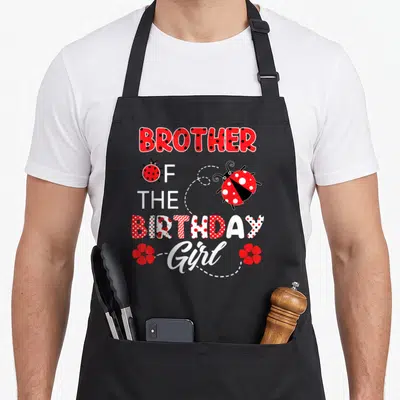 Brother ladybug apron - Stacked BROTHER OF THE BIRTHDAY Girl text with two ladybugs and red flowers connected — shown on a black cotton canvas apron against a white background