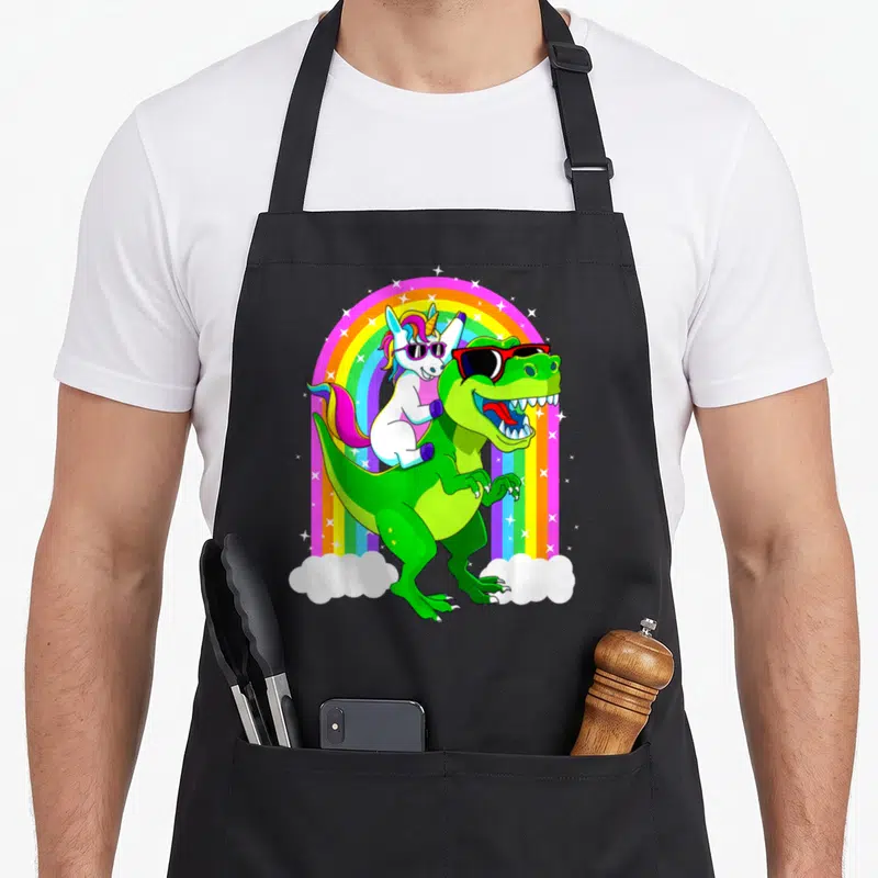 Riding trex dinosaur apron - Cartoon unicorn and t-rex with sunglasses under rainbow arch with stars — shown on a black cotton canvas apron against a white background