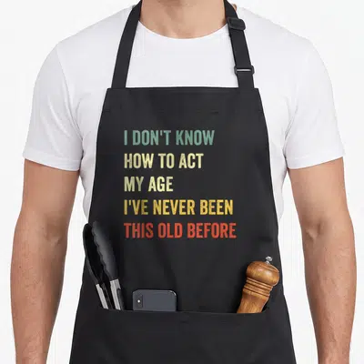 Act age apron for dad - Stacked text reading I DON'T KNOW HOW TO ACT MY AGE in green yellow and red — shown on a black cotton canvas apron against a white background