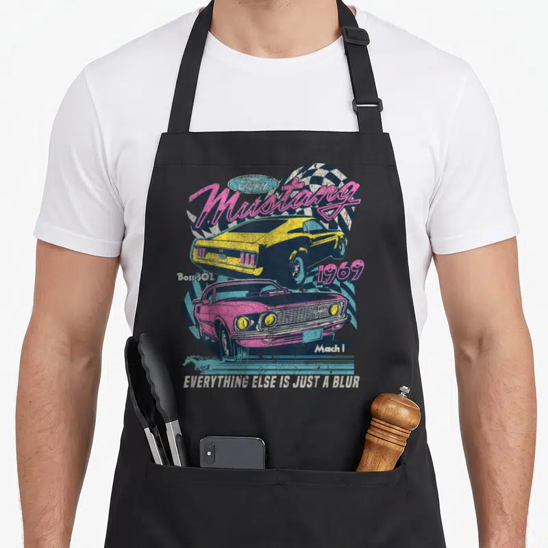 Ford boss apron - Yellow and pink 1969 Ford Mustang cars with Boss 302 text and checkered flag background — shown on a black cotton canvas apron against a white background