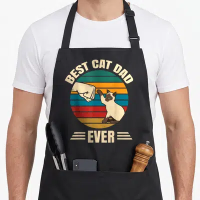 Cool siamese cute cat aprons - Retro fist bump graphic with BEST CAT DAD EVER text and striped circle — shown on a black cotton canvas apron against a white background