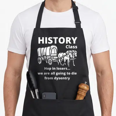 History sarcastic apron - White covered wagon with two horses and text Hop in losers we are all going to die — shown on a black cotton canvas apron against a white background