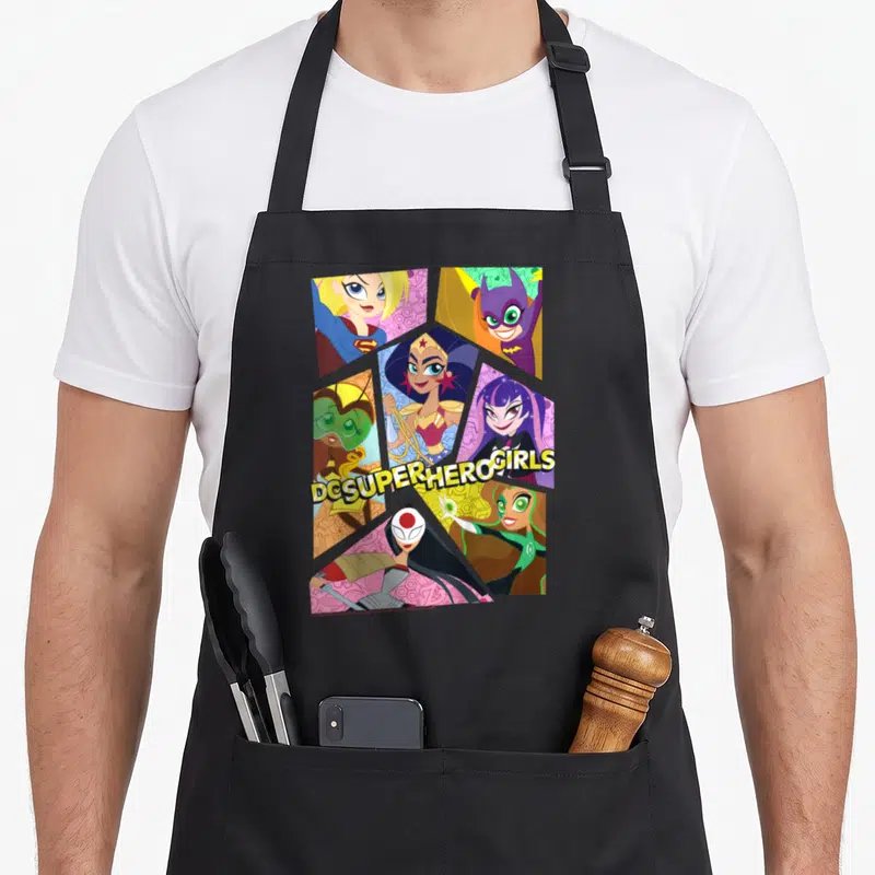 Collage super hero aprons - Six cartoon superhero girls in honeycomb layout with DC SUPER HERO GIRLS text — shown on a black cotton canvas apron against a white background
