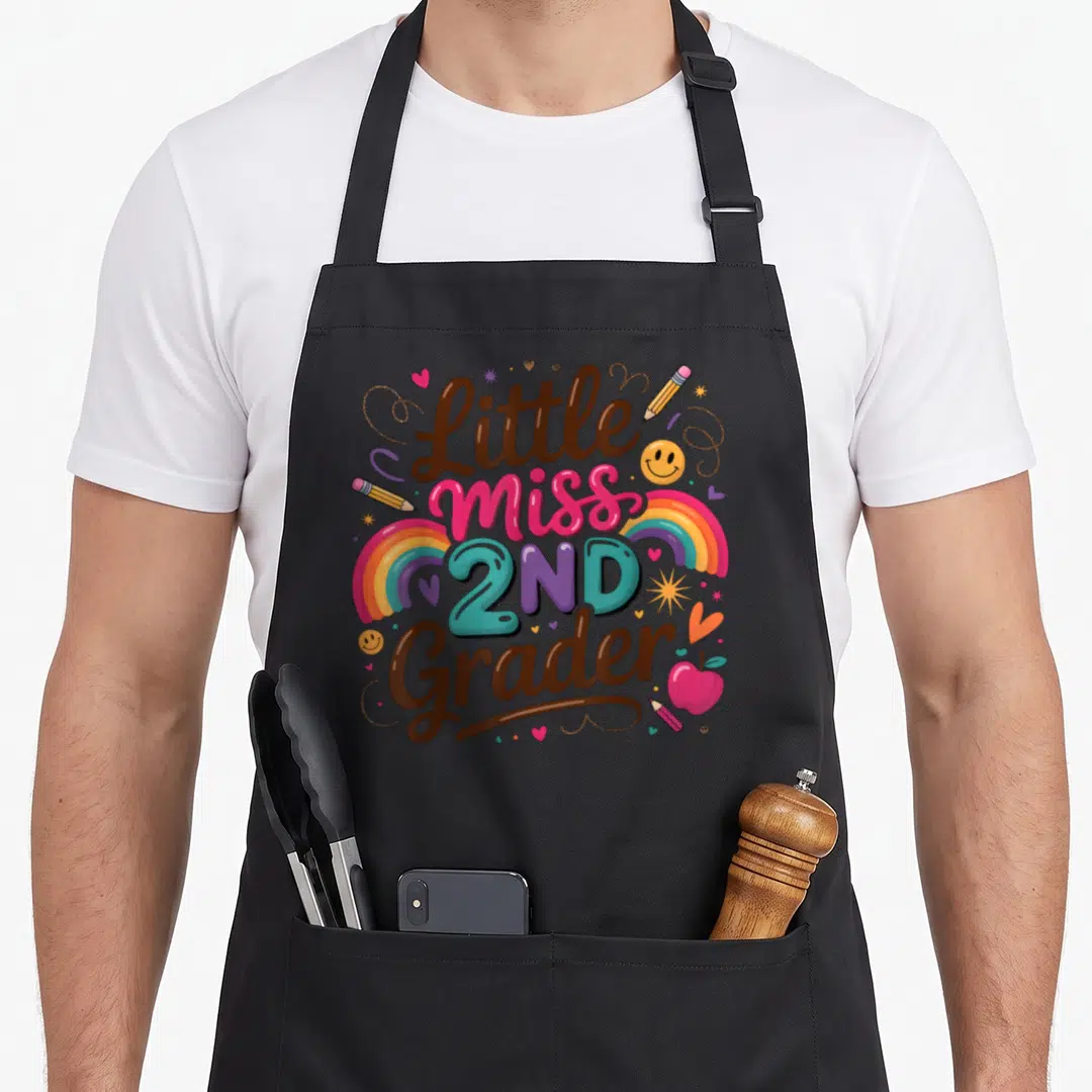 Grade second 2nd apron - Little Miss 2ND Grader text with rainbows, pencils, hearts, and smiley faces in brown and pink — shown on a black cotton canvas apron against a white background