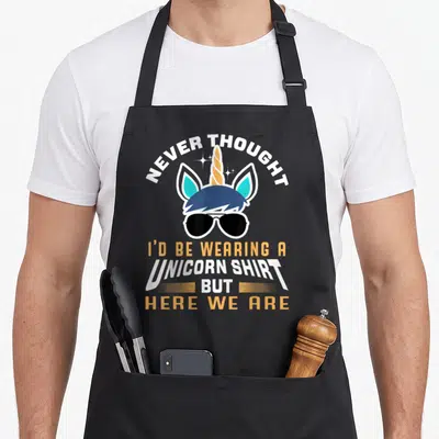 Unicorn grandpa apron - Cartoon unicorn head with sunglasses and arched text reading Never Thought I'd Be Wearing — shown on a black cotton canvas apron against a white background