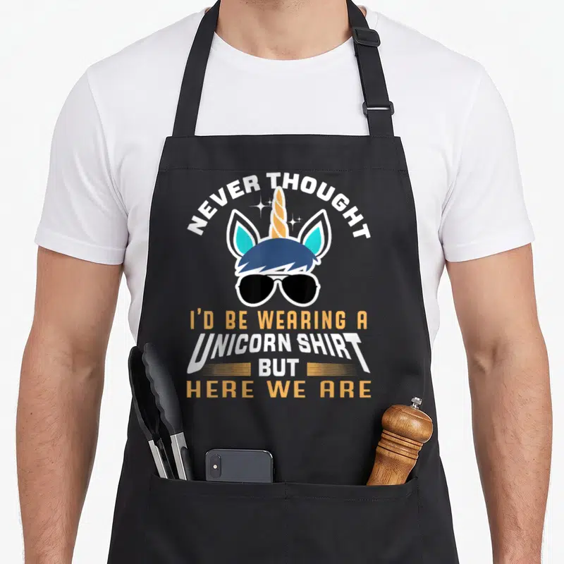 Unicorn grandpa apron - Cartoon unicorn head with sunglasses and arched text reading Never Thought I'd Be Wearing — shown on a black cotton canvas apron against a white background