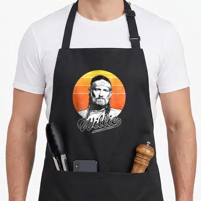 Willie sunset apron - Distressed portrait of a man with braids against a striped orange and yellow sun with Willie — shown on a black cotton canvas apron against a white background