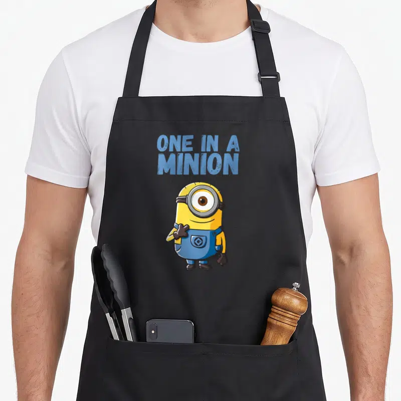 Blue despicable me apron - ONE IN A MINION text above yellow minion standing in blue overalls — shown on a black cotton canvas apron against a white background