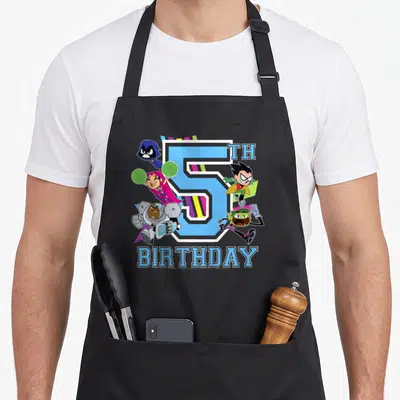 Funny 5th birthday apron - Blue number 5 with five cartoon characters and diagonal stripes in green purple pink — shown on a black cotton canvas apron against a white background