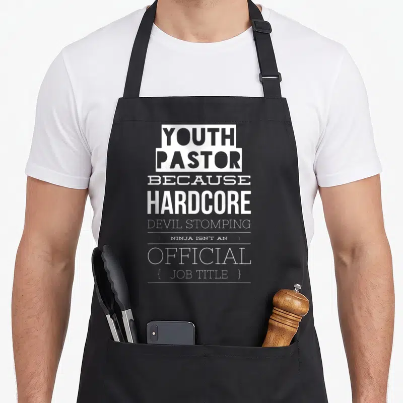 Youth devil's apron - White stacked text YOUTH PASTOR BECAUSE HARDCORE DEVIL STOMPING Ninja Official Job Title — shown on a black cotton canvas apron against a white background