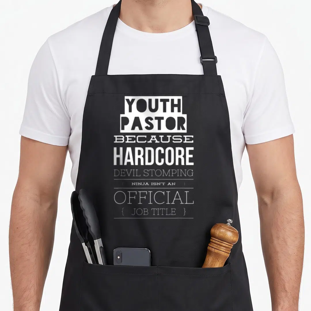 Youth devil's apron - White stacked text YOUTH PASTOR BECAUSE HARDCORE DEVIL STOMPING Ninja Official Job Title — shown on a black cotton canvas apron against a white background