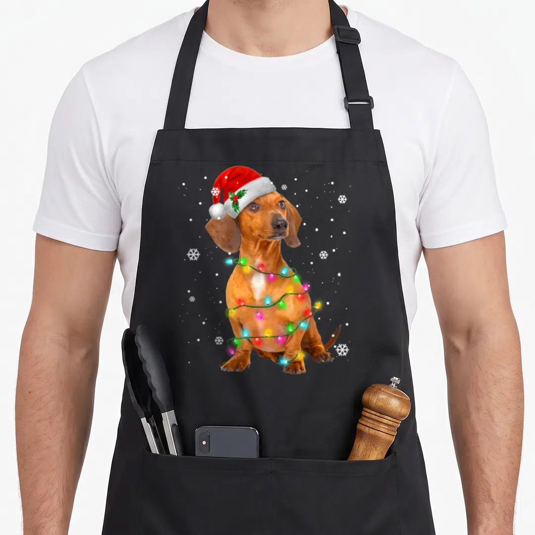 Tree funny dog apron - Dachshund in red santa hat wrapped in colorful christmas lights surrounded by white snowflakes — shown on a black cotton canvas apron against a white background
