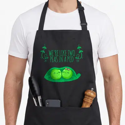 Pod couple apron - Cartoon green peas in pod with WE'RE LIKE TWO PEAS IN A POD text and pink bow — shown on a black cotton canvas apron against a white background