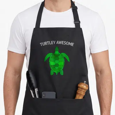 Turtley joke apron - Green cartoon turtle below white arched text reading TURTLEY AWESOME — shown on a black cotton canvas apron against a white background