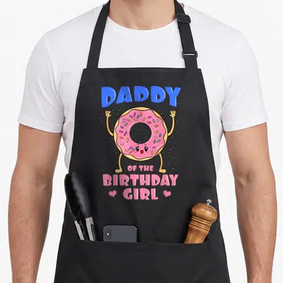Daddy donut apron - Cartoon tan donut with pink frosting and blue DADDY text flanked by two hearts — shown on a black cotton canvas apron against a white background