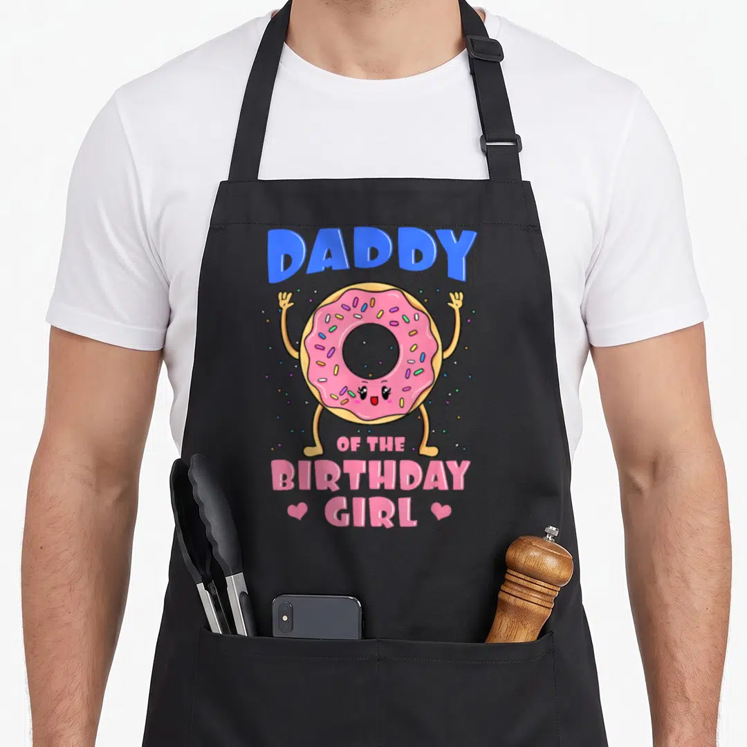 Daddy donut apron - Cartoon tan donut with pink frosting and blue DADDY text flanked by two hearts — shown on a black cotton canvas apron against a white background