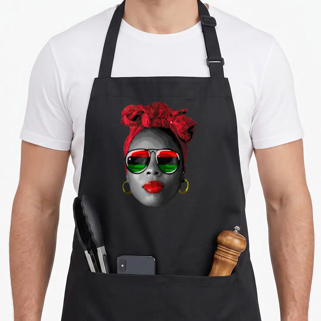Juneteenth chef outfit apron - Woman portrait with red headwrap, gold hoops, and sunglasses reflecting red black green — shown on a black cotton canvas apron against a white background