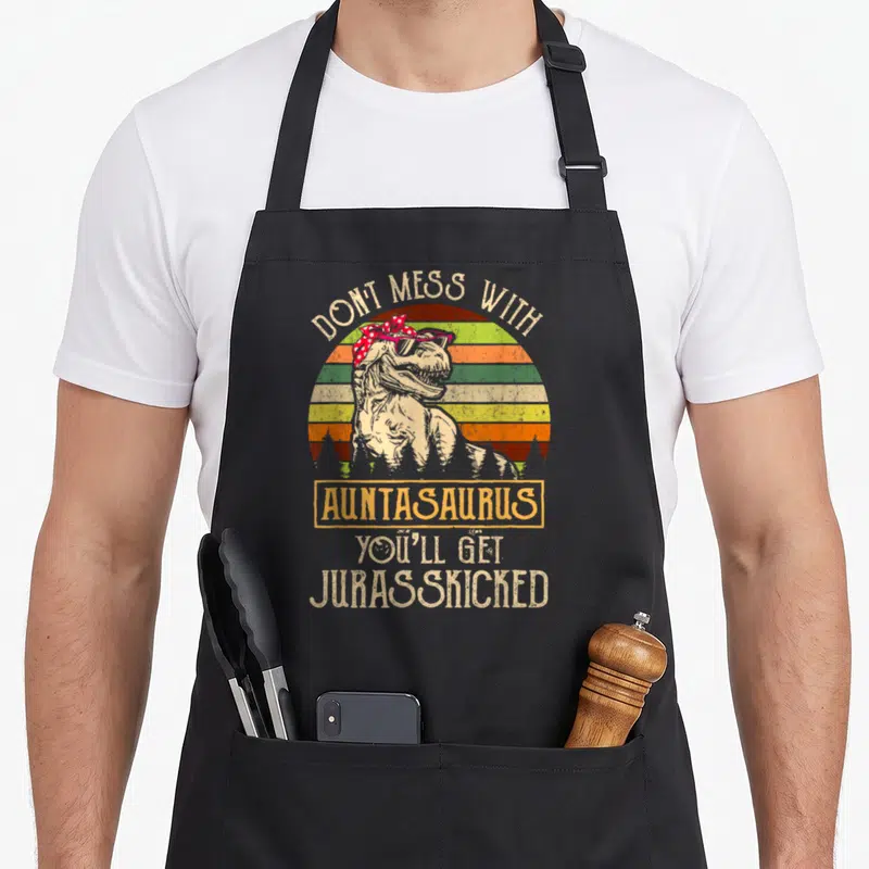 Mess dinosaur apron - Retro T-Rex in sunglasses with striped sun and 'Don't Mess With Auntasaurus' text — shown on a black cotton canvas apron against a white background
