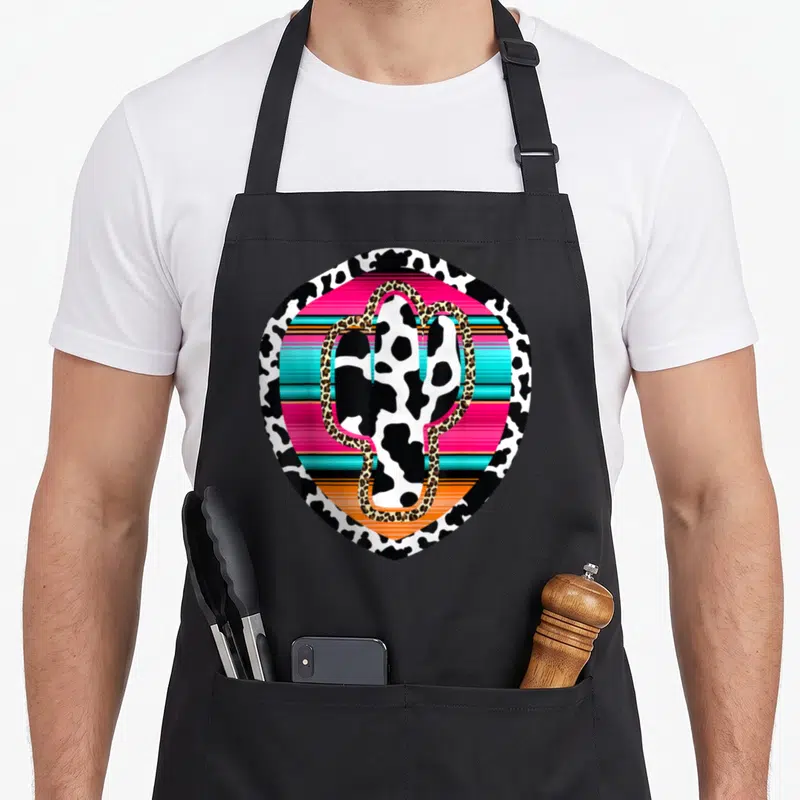 Western cow apron - Cow-print cactus with leopard outline over pink, turquoise, and orange stripes in a shield shape — shown on a black cotton canvas apron against a white background