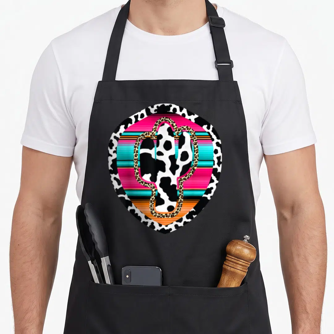 Western cow apron - Cow-print cactus with leopard outline over pink, turquoise, and orange stripes in a shield shape — shown on a black cotton canvas apron against a white background