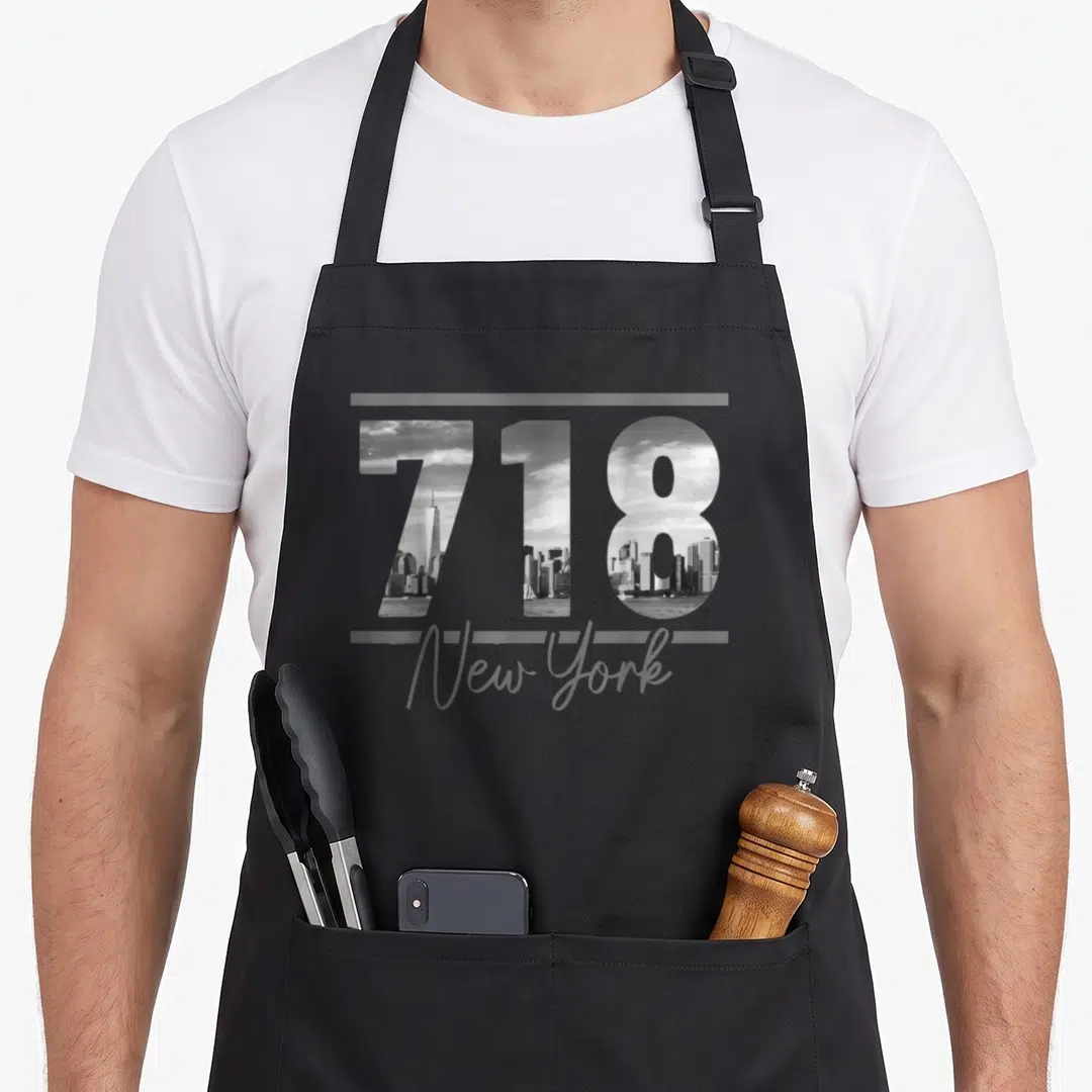 Code new york aprons - 718 number filled with cityscape and New York script in grey and black — shown on a black cotton canvas apron against a white background